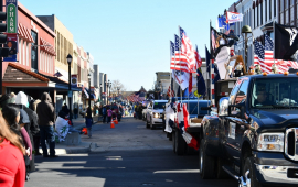 Vehicles with American flags drive in a parade down a street lined with people waving and historic buildings