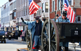 Man in Union Civil War replica uniform rides on a parade float with an American flag and waves. Behind him are historic building