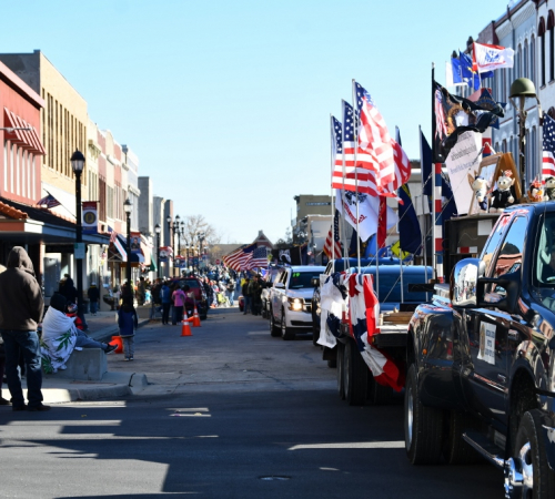 Vehicles with American flags drive in a parade down a street lined with people waving and historic buildings