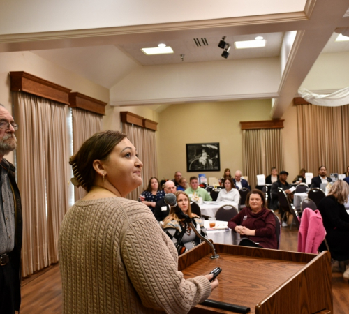 man and woman speak in front of podium to address audience seated at tables inside room