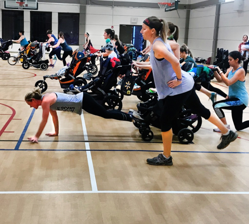 A group of moms running and walking with their strollers during the class in the Riverfront Community Center gym