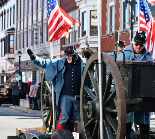 Man in Union Civil War replica uniform rides on a parade float with an American flag and waves. Behind him are historic building
