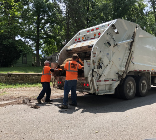 two workers in safety orange shirts load an item into a trash truck