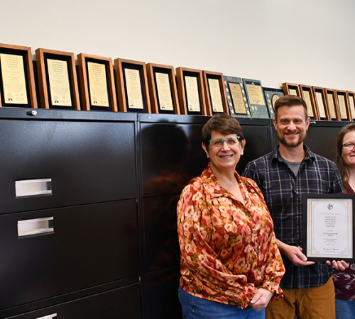 Four people stand in front of filing cabinets with a line of award plaques on top. They are holding another award.