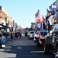 Vehicles with American flags drive in a parade down a street lined with people waving and historic buildings