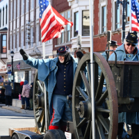 Man in Union Civil War replica uniform rides on a parade float with an American flag and waves. Behind him are historic building