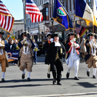 Veterans Day Parade 5 flag bearers in 1700s dress walk down a downtown street in this modern color photo of a Veterans Day Parade