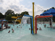 public pool with a large metal water sprinkler feature with dozens of children and families swimming