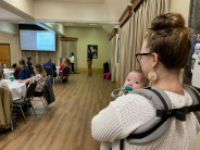 woman holding baby in a harness watches speaker