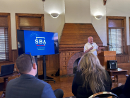 speaker presents to people sitting in chairs, initials S-B-A and Small Business Administration are on screen