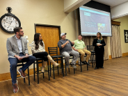 Leavenworth business owner panel addresses audience at Business Symposium Woman in dress pants and blazer stands and holds microphone next to four people seated in high chairs inside building