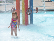 Child stands in shallow water at Wollman Aquatic Center as water falls from overhead features