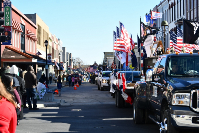 Vehicles with American flags drive in a parade down a street lined with people waving and historic buildings