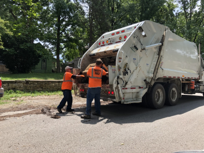 two workers in safety orange shirts load an item into a trash truck