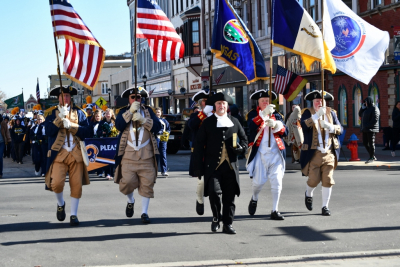 5 flag bearers in 1700s dress walk down a downtown street in this modern color photo of a Veterans Day Parade