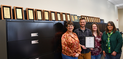 Four people stand in front of filing cabinets with a line of award plaques on top. They are holding another award.