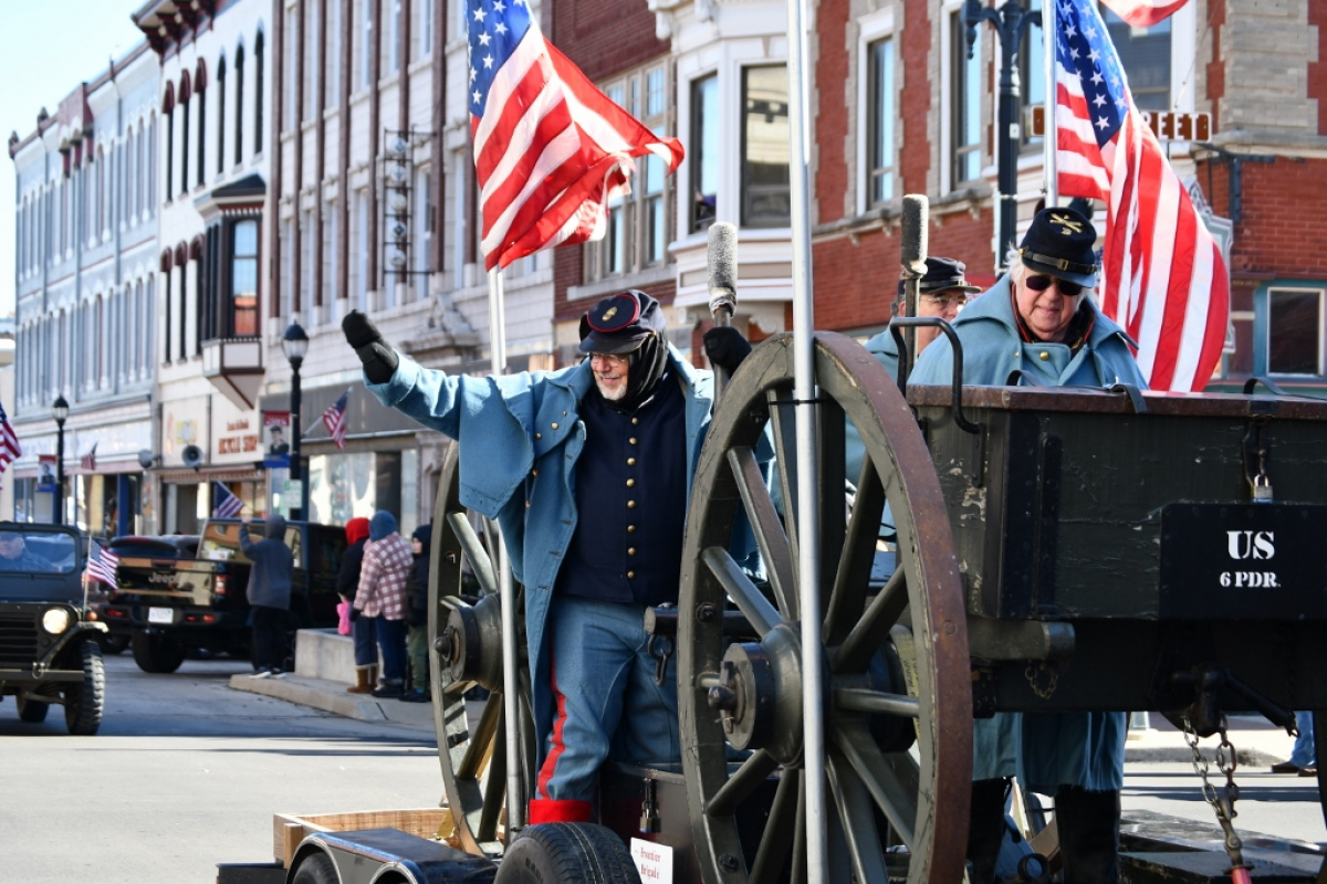 Veterans Day Parade Man in Union Civil War replica uniform rides on a parade float with an American flag and waves. Behind him are historic building