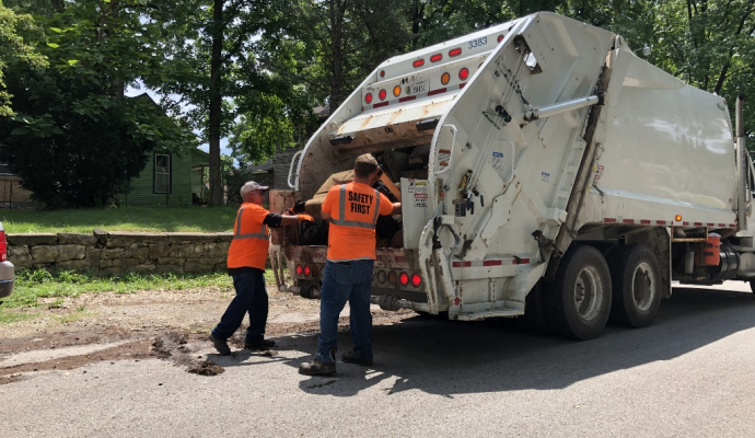 two workers in safety orange shirts load an item into a trash truck