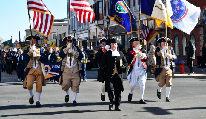 5 flag bearers in 1700s dress walk down a downtown street in this modern color photo of a Veterans Day Parade